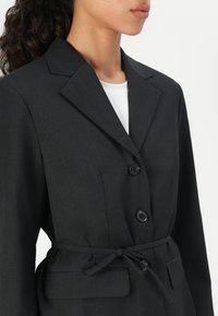 Sacou negru croit, cu un design slim, rever mari, trei butoane și o centură la talie. Material neted, texturat.