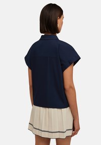 Camicia blu navy a maniche corte con colletto, caratterizzata da un fit rilassato. Abbinata a una gonna plissettata crema con dettagli in blu navy.