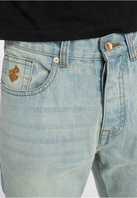 Heledad jeans helesin sinise värviga, millel on kulunud pesu, kontrastsete pruunide õmblustega ja logo detail tagakotis. Hõbedane nööp sulgemiseks.
