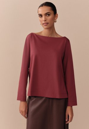 Kvinde med mørkt hår i en lav knold iført en bordeaux langærmet top og en mørkebrun nederdel mod en beige baggrund.