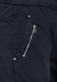 Laadunmukaiset tummansiniset kangashousut sisältävät etutaskun, jossa on hopeinen vetoketju ja näkyvä neppari. Kankaan pinta vaikuttaa sileältä, ja siinä on hienovaraisia ompelutaitoja.