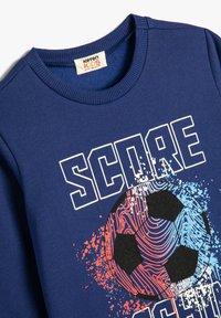 Sudadera azul con cuello redondo y puños de canalé. El gráfico frontal incluye la palabra "SCORE" y un balón de fútbol negro estilizado con salpicaduras de colores.