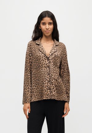 Femme portant une chemise à boutons à imprimé léopard marron et noir avec un col et un pantalon noir, debout devant un fond clair uni.