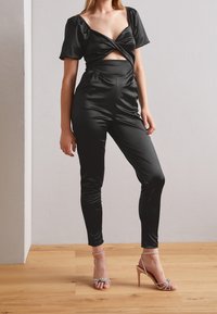 Mono de satén negro con mangas cortas abullonadas y detalle cortado en la cintura. Combinado con sandalias de tacón alto metálicas con tiras.