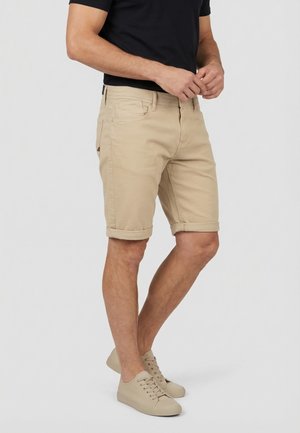 Uomo con pantaloncini beige risvoltati, scarpe da ginnastica beige e maglietta nera, in piedi con le mani unite davanti.