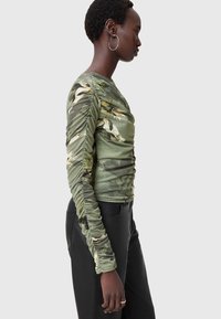 Langärmliges Shirt in Olivgrün mit Blumenmuster, ausgestattet mit Raffung an den Ärmeln und tailliertem Schnitt. Kombiniert mit schwarzen Hosen.