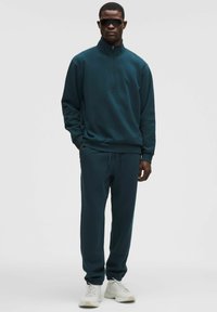 Sudadera de cremallera en verde azulado oscuro con cuello alto y pantalones jogger a juego, confeccionados en una tela suave. Combinada con zapatillas de color claro.