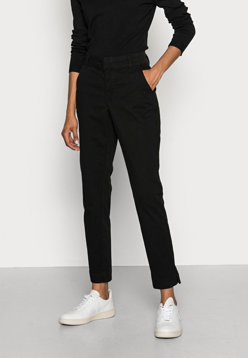 Personne portant un pantalon noir taille haute fuselé et un haut noir à manches longues, une main dans la poche, et des baskets blanches sur un sol gris.