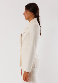 Blazer su misura beige chiaro con design strutturato, rever a punta, maniche lunghe e tasche laterali. Texture del tessuto liscia.