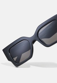 Black oversized sunglasses with a thick matte frame. Dark gradient lenses, angular shape, and minimal branding inside the frame.