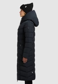 Palton puffer negru, până la genunchi, cu glugă, având un model matlasat în formă de zigzag și închidere cu fermoar. Însoțit de o căciulă tricotată neagră.