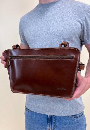 Brown leather bag with a smooth finish, one front zipper pocket, double stitching, and adjustable straps. Held by a person.