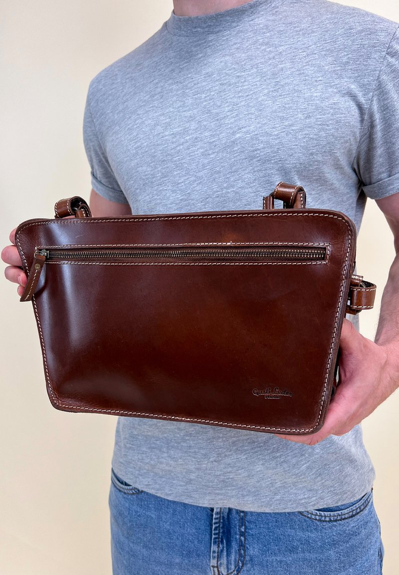 Bruine leren tas met een gladde afwerking, één voorzak met rits, dubbele stitching en verstelbare banden. Vastgehouden door een persoon.