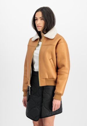 Woman wearing a tan shearling collar jacket, white top, and black quilted button-front skirt, standing against a white background.