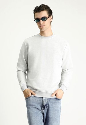 Lichtgrijze sweatshirt met een ronde halslijn en ribgebreide manchetten, gecombineerd met blauwe jeans. Model draagt donkere, rechthoekige zonnebril.