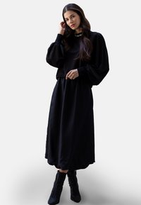 Schwarzes Langarmshirt mit gepufften Ärmeln, kombiniert mit einem langen schwarzen Rock und schwarzen, verzierten Stiefeletten. Weicher Stoff mit glatter Textur.