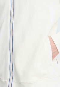 Sweat à capuche blanc à fermeture éclair, fabriqué dans un tissu texturé, avec une fermeture éclair bleue contrastante et des poches latérales. Détail de couture remarquable le long de l'ourlet.