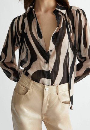 Kobieta w czarno-beżowej bluzce w abstrakcyjne paski, włożonej do wysokowysłanych beżowych skórzanych spodni.