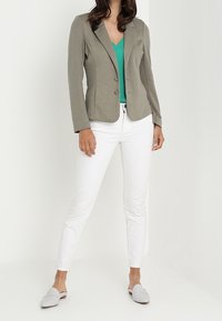 Harmaa istuva blazer, jossa on kaksi nappia, yhdistetty vihreään puseroon. Valkoiset slim-fit-housut ja vaaleanharmaat liukujalkineet. Tasaiset tekstuurit kauttaaltaan.