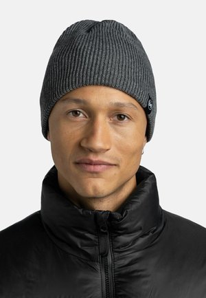 Gray ribbed knit beanie with a slightly slouchy shape. Features a black logo patch on the side. Worn with a black zippered puffer jacket.