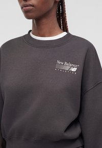 Άτομο που φοράει σκούρο γκρι φούτερ New Balance Athletics με λευκό λογότυπο στο στήθος, εμφανή κοτσιδάκια, απλό φωτεινό φόντο.