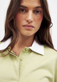 Hellgrünes Poloshirt mit einem weißen Kragen, ausgestattet mit einer Dreiknopfleiste und geripptem Textil, teilweise unter dem Gesicht sichtbar.