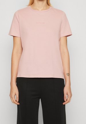 Femme portant un t-shirt uni rose clair à manches courtes et un pantalon noir, avec un petit tatouage en forme de cœur sur son poignet gauche.