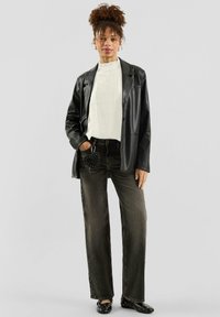 Schwarzer Lederblazer, weißer gerippter Pullover, dunkle Jeans, herzförmiges Accessoire am Gürtel, schwarze Nieten-Ballerinas, stehende Pose, neutraler Hintergrund.