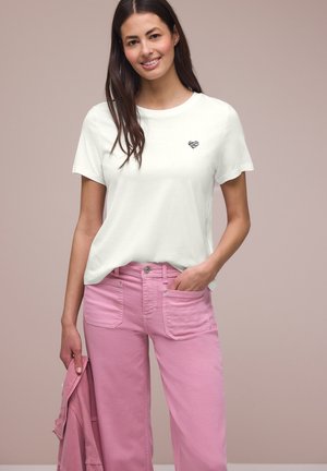 Frau lächelt, trägt ein weißes T-Shirt mit kleinem Herz-Logo und rosa weite Hosen, hält eine passende rosa Jacke.