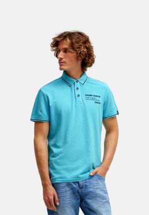 Junger Mann mit lockigem Haar, der ein blau gestreiftes Poloshirt mit dem Aufdruck "CAMP DAVID" und blaue Jeans trägt, schaut nach links, eine Hand in der Tasche.