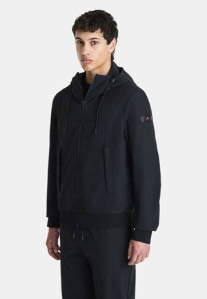 Giovane uomo che indossa una giacca nera con cappuccio e cerniera, con tasche laterali e polsini a coste, in piedi davanti a uno sfondo chiaro e uniforme.