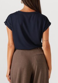 Top de manga corta en color navy con puños enrollados, combinado con pantalones marrones ajustados que cuentan con un bolsillo trasero y detalle de cremallera discreto.