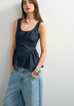 Kvinde iført en ærmeløs mørk denim peplum-top, lyseblå løstsiddende jeans og metalliske armbånd med hænderne i lommerne mod en hvid væg.