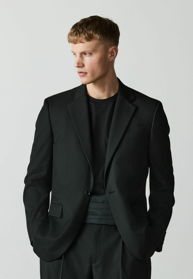 Jeune homme aux cheveux courts portant un blazer noir, une chemise noire et un pantalon noir, regardant vers sa gauche sur un fond clair uni.