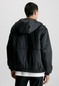 Giacca nera imbottita con cappuccio, orlo e polsini elasticizzati. Realizzata in materiale leggero e liscio, presenta un design semplice senza decorazioni.