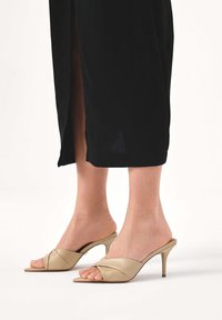 Beżowe skórzane klapki na obcasie z otwartym palcem, z ozdobnym skręcanym wzorem na cholewce, niskim obcasem typu szpilka oraz gładką fakturą.