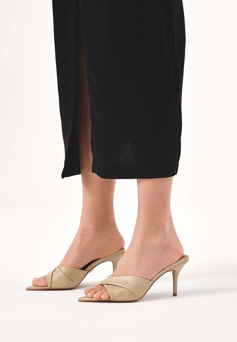 Beżowe skórzane klapki na obcasie z otwartym palcem, z ozdobnym skręcanym wzorem na cholewce, niskim obcasem typu szpilka oraz gładką fakturą.