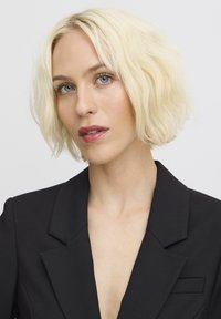 Krátký černý blazer s hlubokým véčkovým výstřihem a decentními klopami. Model má vlnité platinově blond vlasy a modré oči, na bílém pozadí.