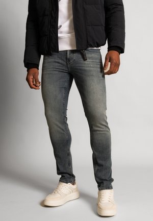 Mężczyzna ubrany w wyblakłe szare wąskie dżinsy, białe tenisówki, białą bluzę oraz czarną puchową kurtkę, stojący na tle szarego tła.