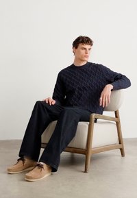 Granatowy, teksturowany sweter, czarne spodnie o szerokich nogawkach i beżowe zamszowe buty. Siedzący na jasnym fotelu z drewnianą ramą. Minimalistyczne tło.