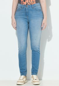 Donna che indossa jeans skinny azzurri chiari, sneakers beige e bianche, e una maglietta nera con testo arancione, in piedi su uno sfondo bianco.