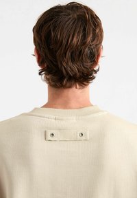 Felpa beige con colletto a coste, cuciture in vista e una toppa rettangolare in tessuto con due occhielli metallici sul retro.