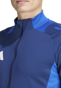 Giacca sportiva blu navy con colletto alto, chiusura a zip e dettagli a contrasto azzurro chiaro sulle spalle e ai lati. Dettaglio del logo bianco.