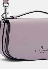 Fioletowa skórzana torebka na ramię z teksturowanym wykończeniem, czarnym obszyciem, regulowanym paskiem i wytłoczonym logo marki z przodu.