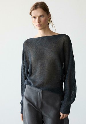 Frau trägt einen transparenten schwarzen Pullover mit langen Ärmeln und U-Boot-Ausschnitt, dazu dunkelgraue Hosen und große goldene Ohrringe vor schlichten Hintergrund.