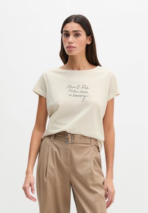 Femme aux longs cheveux foncés portant un t-shirt crème avec un texte et un pantalon beige taille haute avec ceinture, debout devant un fond uni.