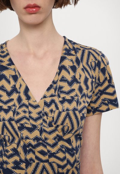 Top a maniche corte in beige con un motivo geometrico blu. Presenta un design a scollatura a V e una texture morbida, che evidenzia forme e linee intricate.