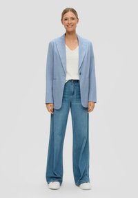 Lichtblauwe blazer over een witte V-hals t-shirt, gecombineerd met wijde blauwe jeans en witte sneakers, tegen een neutrale achtergrond.