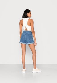 Λευκό αμάνικο crop top με κορδόνια στην πλάτη, συνδυασμένο με μπλε denim σορτς με γυρισμένα στριφώματα και δύο πίσω τσέπες, φορεμένο με ανοιχτόχρωμα sneakers.