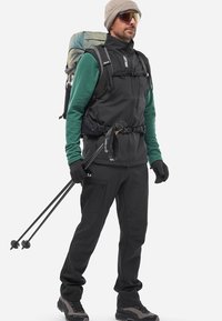 Schwarze Softshell-Weste und Hose, darunter ein grünes Langarmshirt, ausgestattet mit Handschuhen, Trekkingstöcken und einem gemusterten Rucksack.